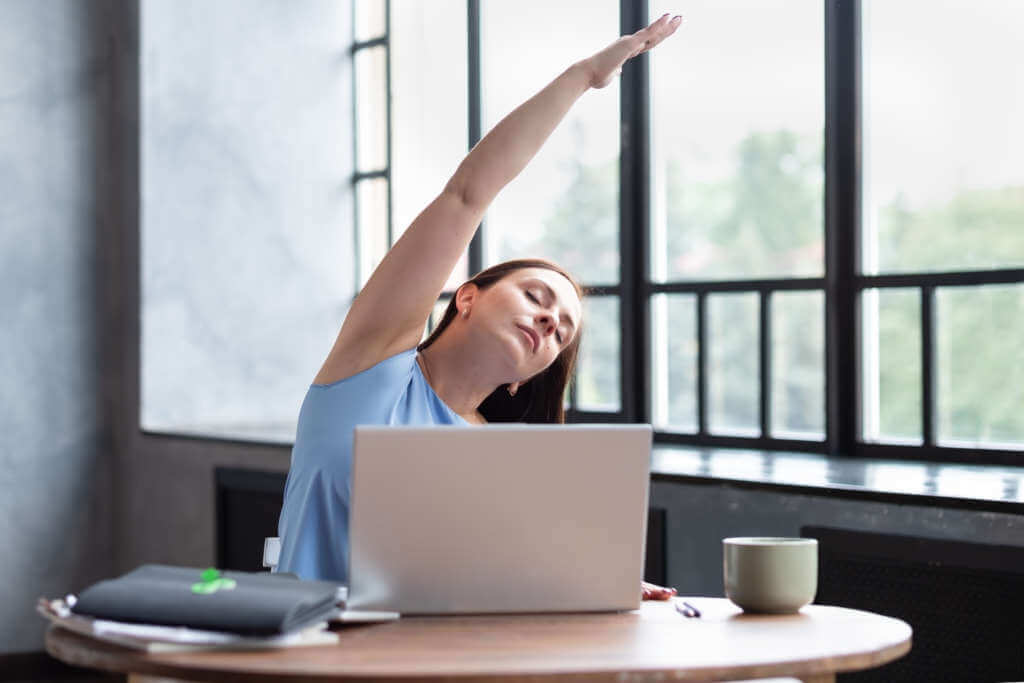 仕事中にノートパソコンの前で腕を伸ばしてストレッチする女性